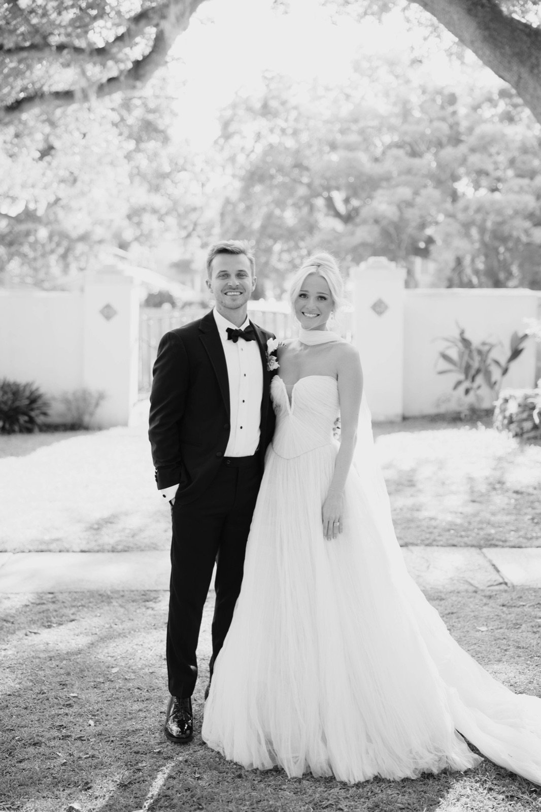 Rick in a tuxedo and his wife in her wedding dress, smiling under an oak tree
