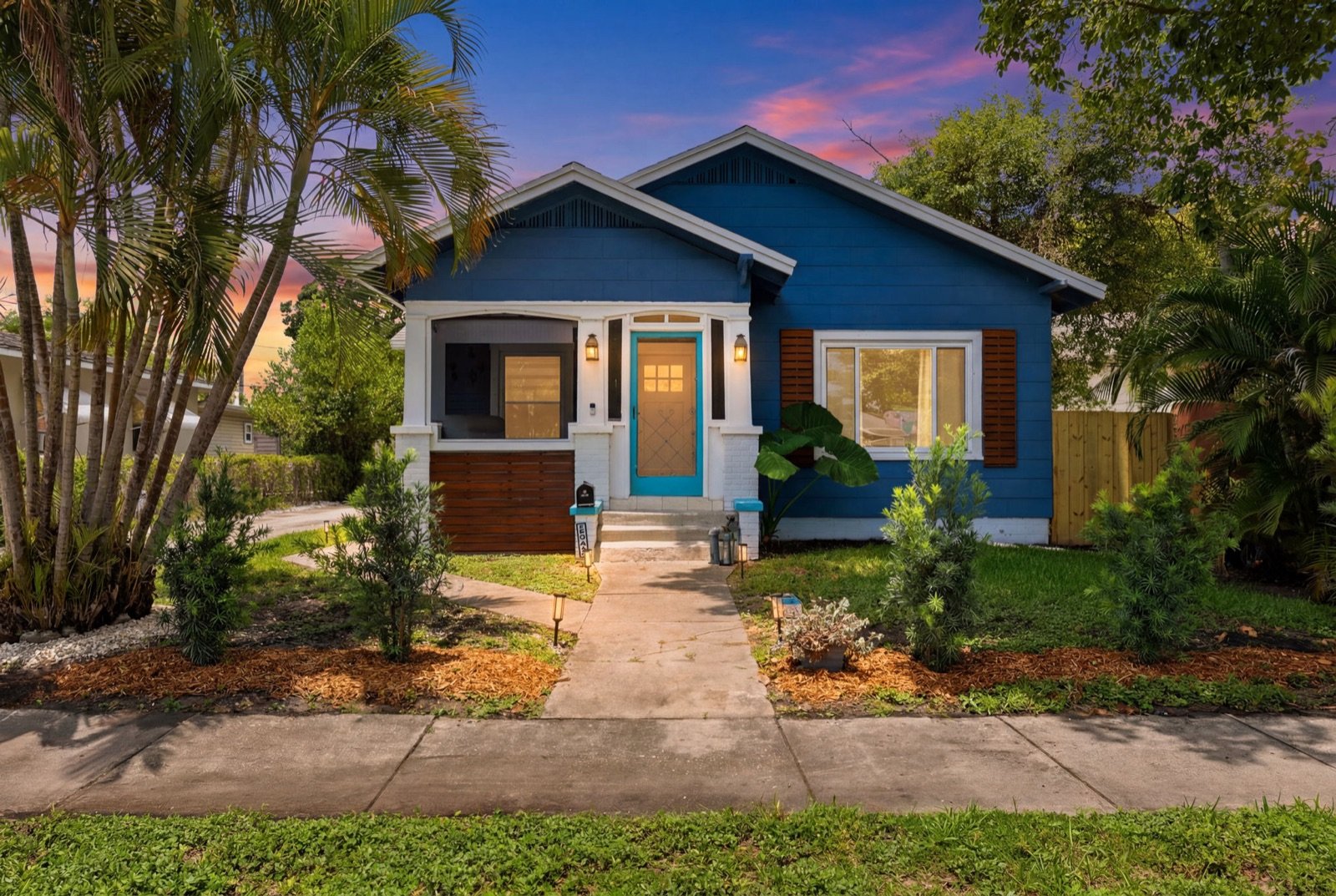 Blue bungalow at twilight with a glowing front door, framed by palms